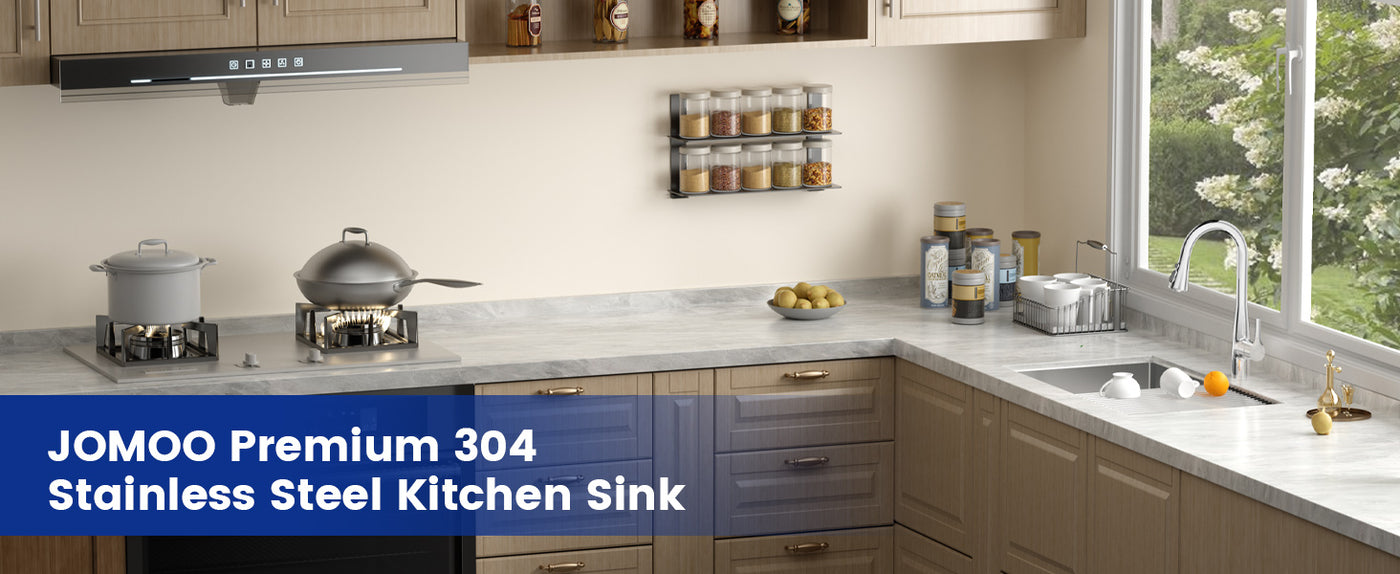 A sleek, contemporary kitchen featuring a JOMOO Premium 304 stainless steel sink, complemented by wooden cabinets, a marble countertop, and a spice rack. The space is adorned with fresh produce and cooking utensils, with natural light streaming through a window overlooking greenery.